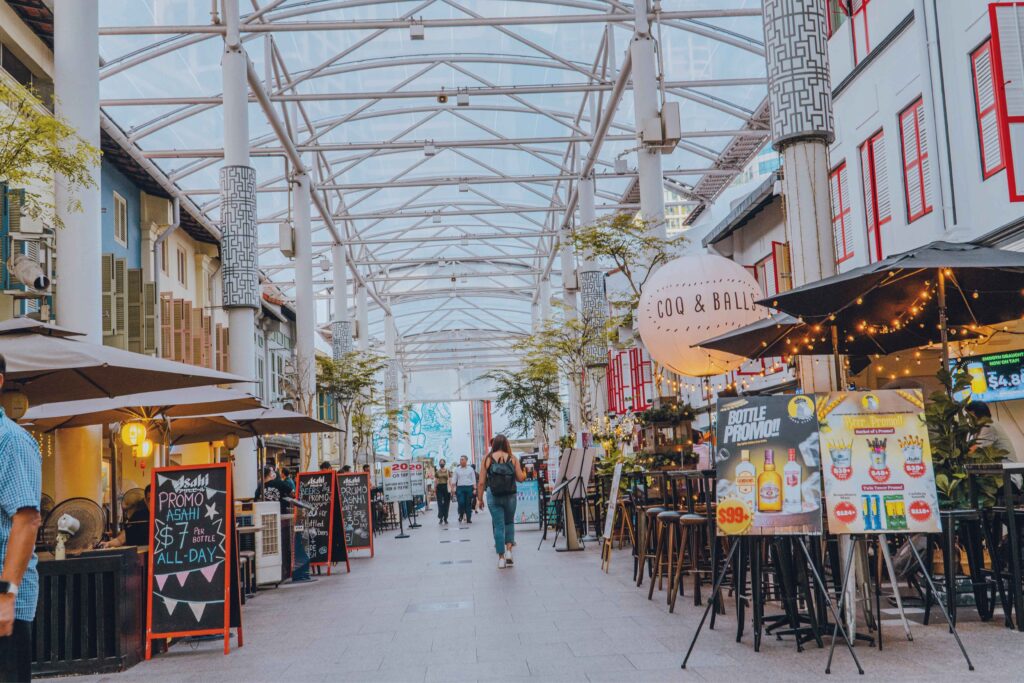 A covered alfresco dining street in Singapore with shophouses, string lights, and people walking past bars and restaurants in the early evening