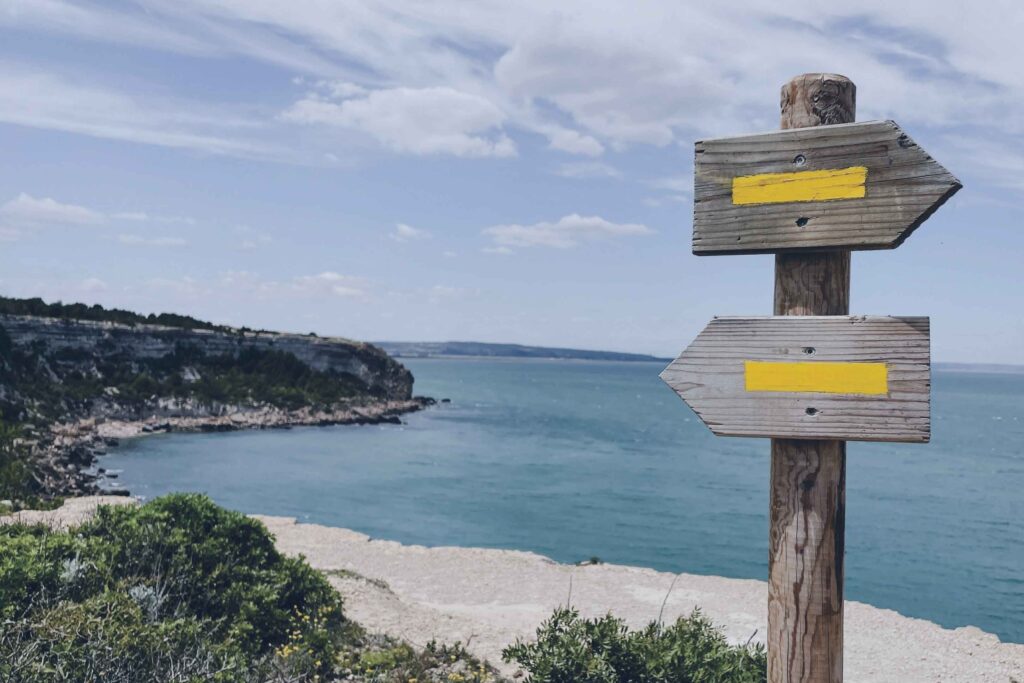 Wooden signpost with two yellow arrows pointing in opposite directions above a coastal bay, representing the retirement trade-off between certainty and higher returns